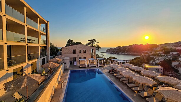 A pool area with lounge chairs and umbrellas, a building with balconies, and a view of the sea and town at sunset.