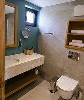 Un baño con lavamanos de mármol, un espejo, una pequeña ventana, un perchero, un cesto de basura y un inodoro.