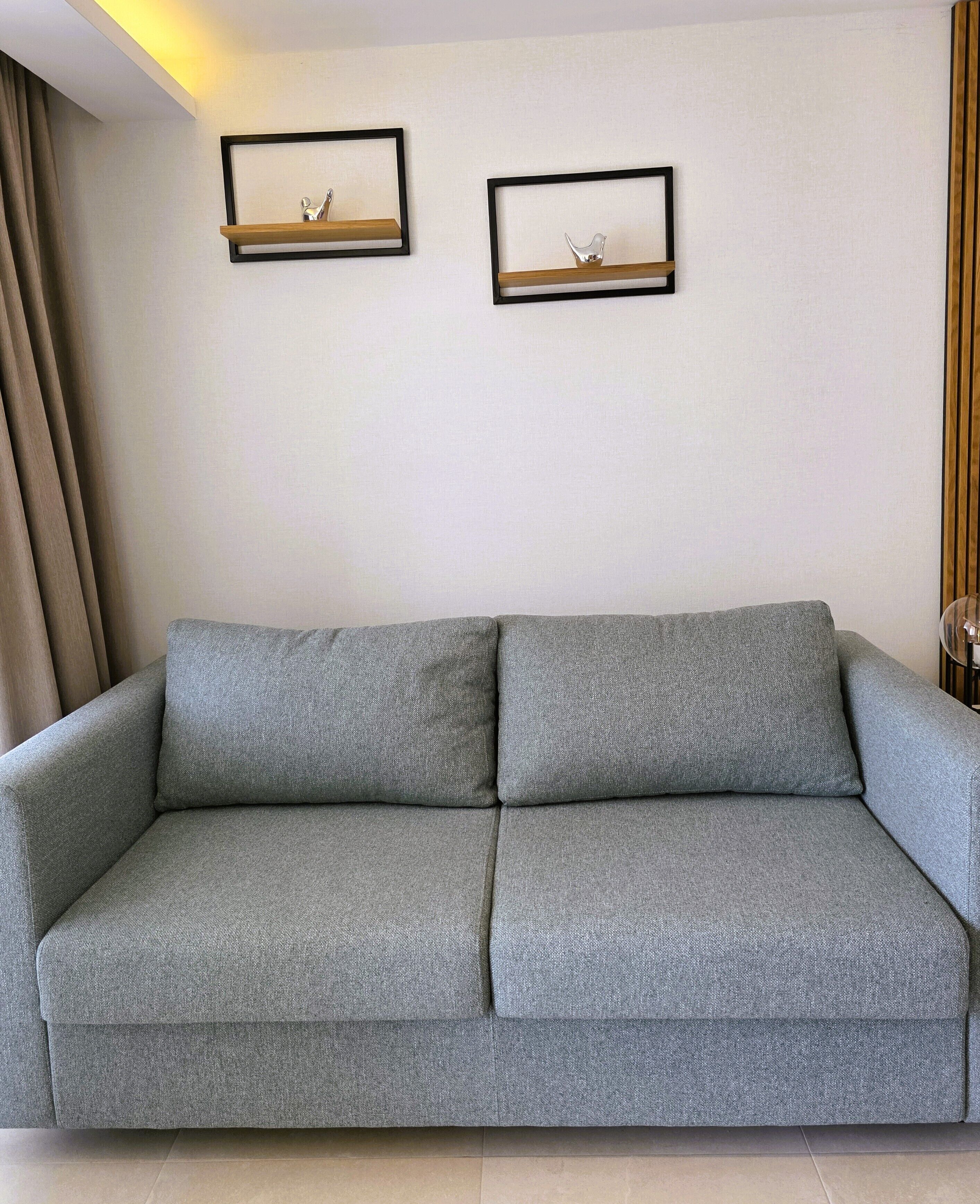 A grey sofa with a wooden frame, two wall-mounted picture frames, and a curtain on the left.