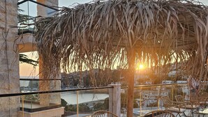 An outdoor seating area with wicker chairs and tables, a thatched umbrella, and a view of the sea.
