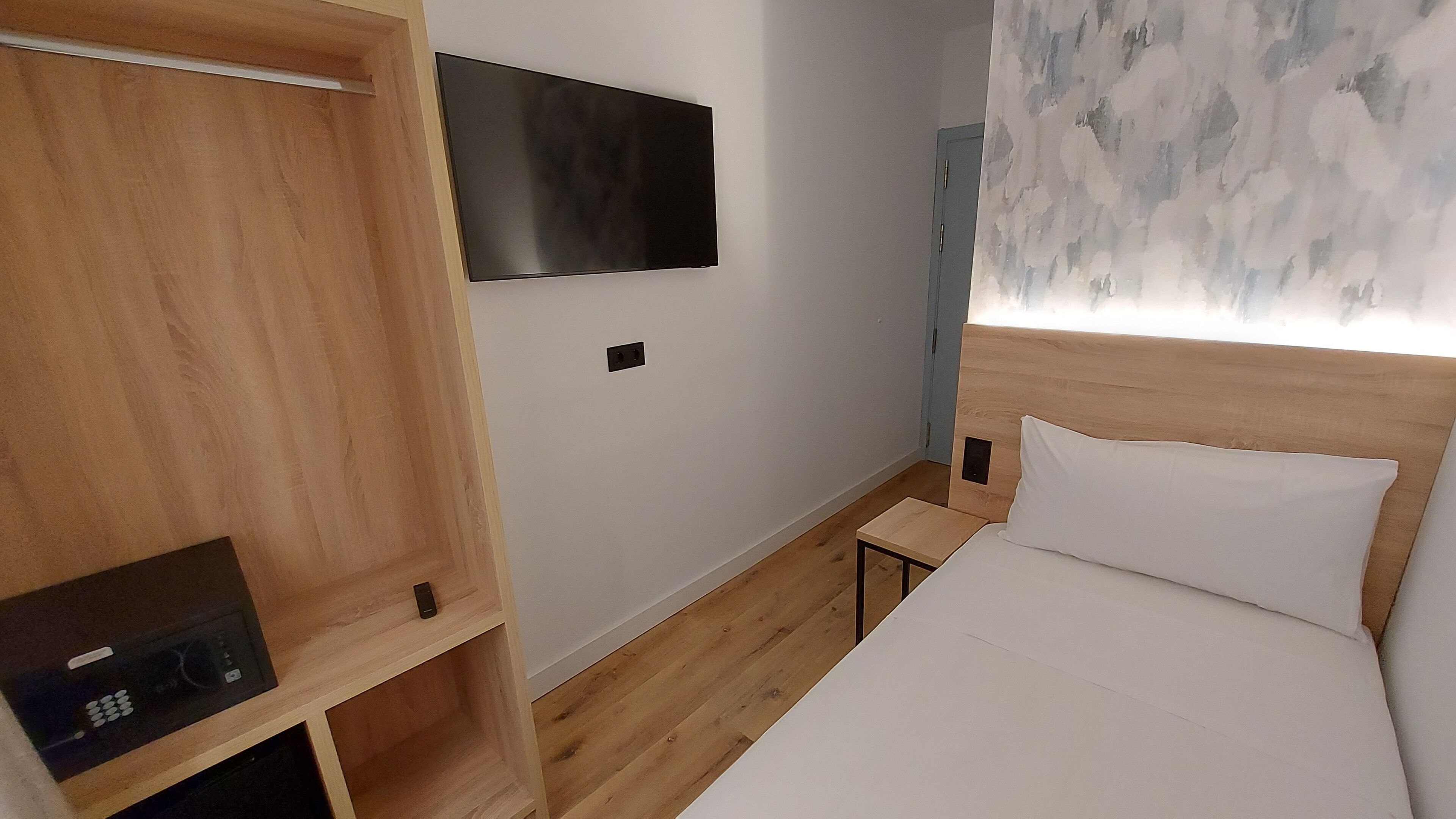 A hotel room with a wooden wardrobe, a flat-screen TV mounted on the wall, a bedside table, and a bed with white bedding.
