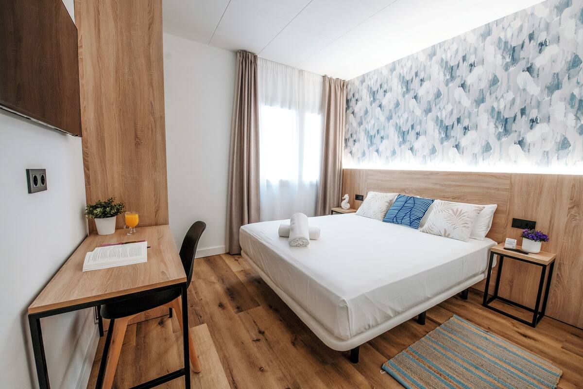 A modern hotel room with a wooden bed, a desk, and a small table with a plant and a glass of orange juice.