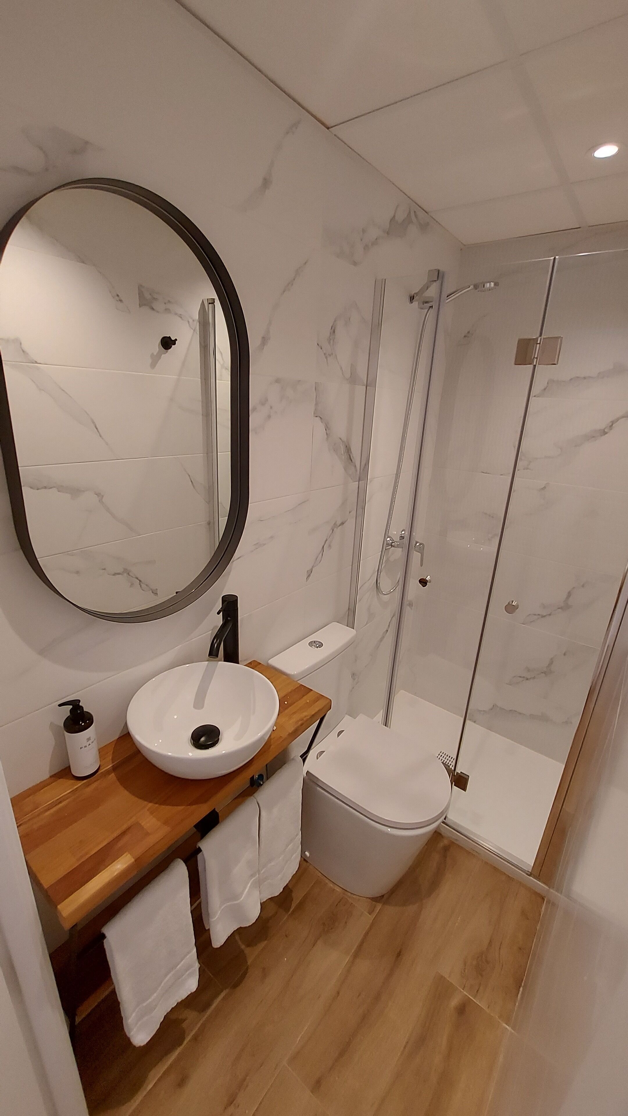 Een moderne badkamer met een marmeren wand, een ronde spiegel, een wastafel, een toilet en een douche met glazen wanden.