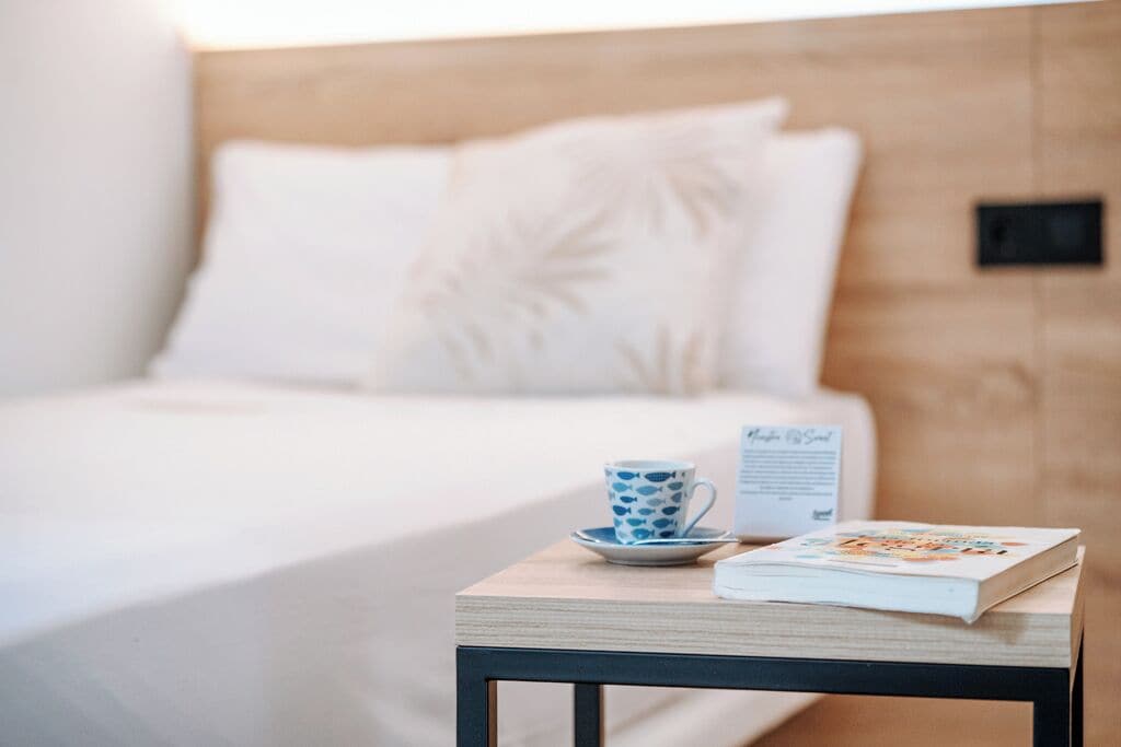 A neatly made bed with a white comforter and a pillow with a leaf pattern. A small table beside the bed holds a blue and white patterned cup, saucer, and a book.