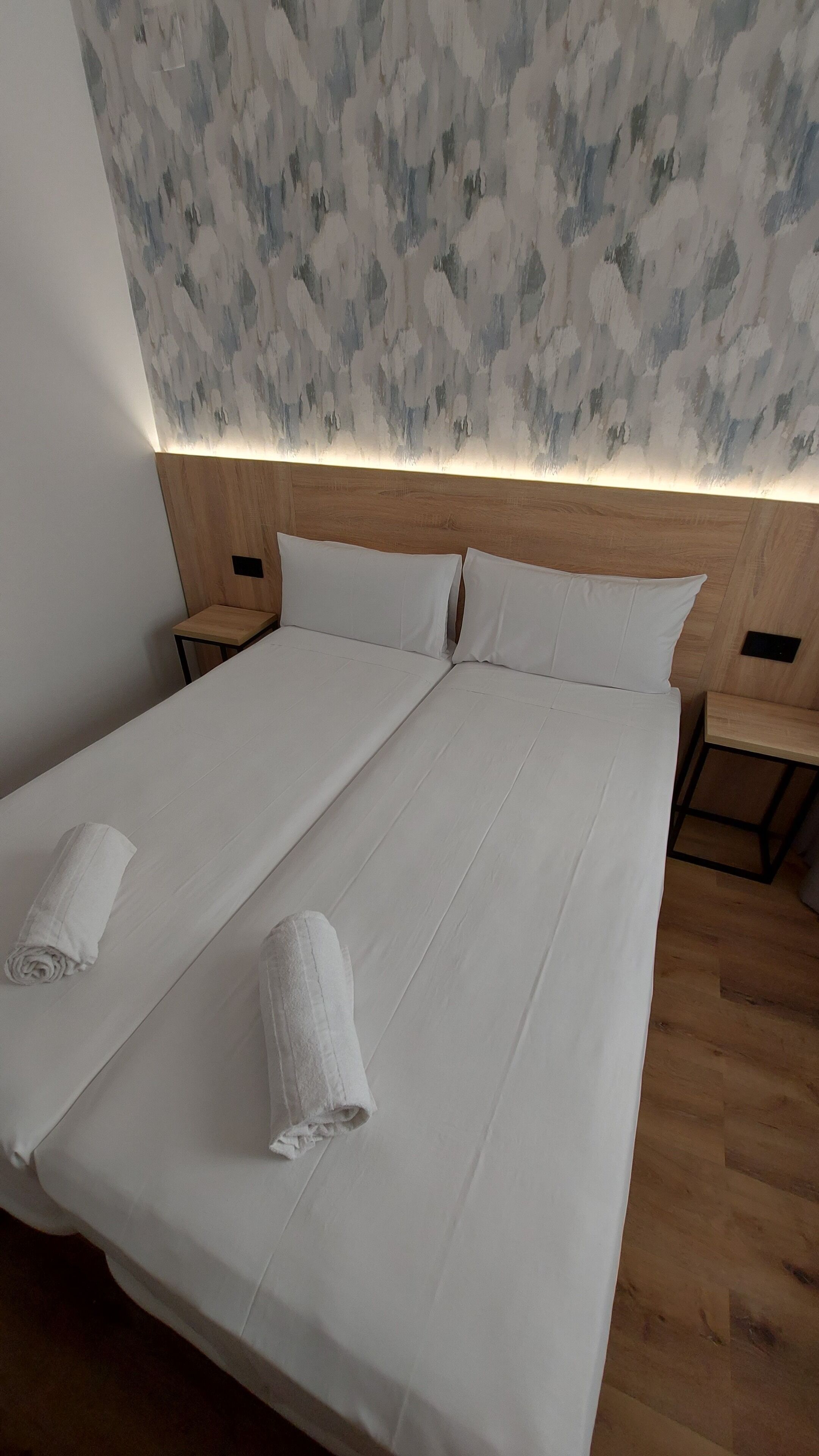 Una cama doble con sábanas blancas y dos toallas enrolladas, cabecera de madera y pared con textura.