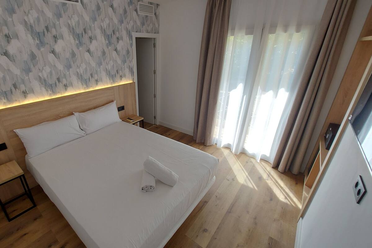 A modern hotel room with a large bed, wooden headboard, and a view of the outdoors through sheer curtains.