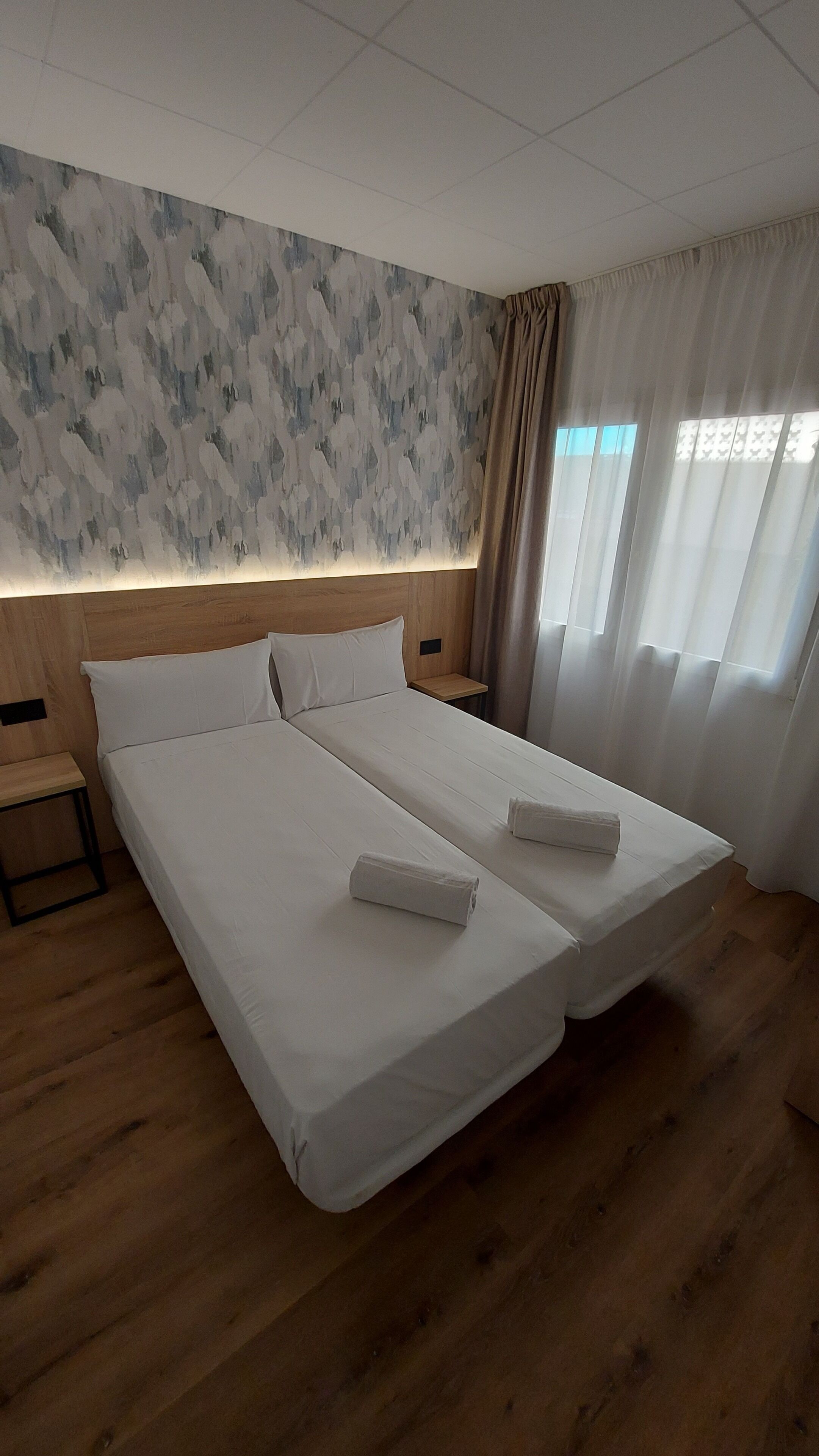 Cama doble con sábanas blancas y dos toallas, cabecera de madera, mesita de noche y ventana con cortinas.