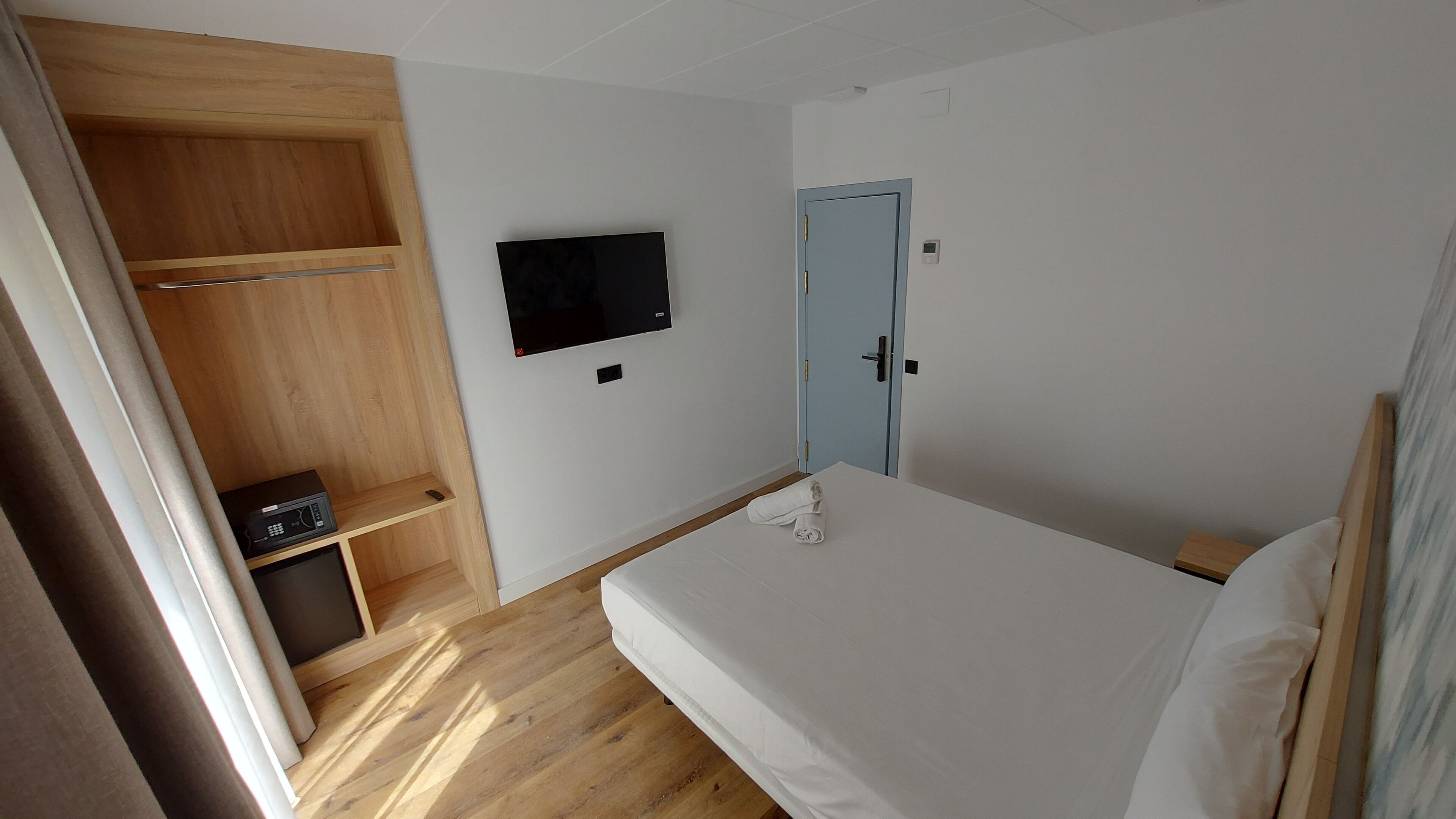A bedroom with a wooden wardrobe, a flat-screen TV, a light blue door, and a bed with white bedding.
