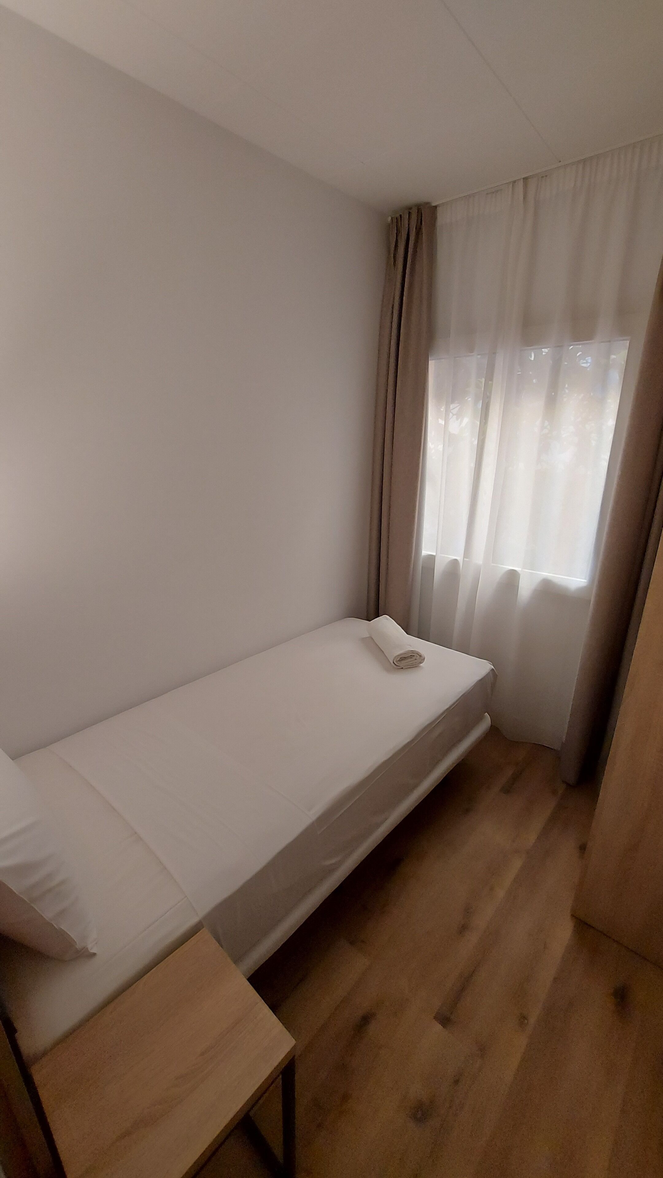Een kleine kamer met een enkel bed, een houten vloer, een klein tafeltje en een raam met dunne gordijnen.