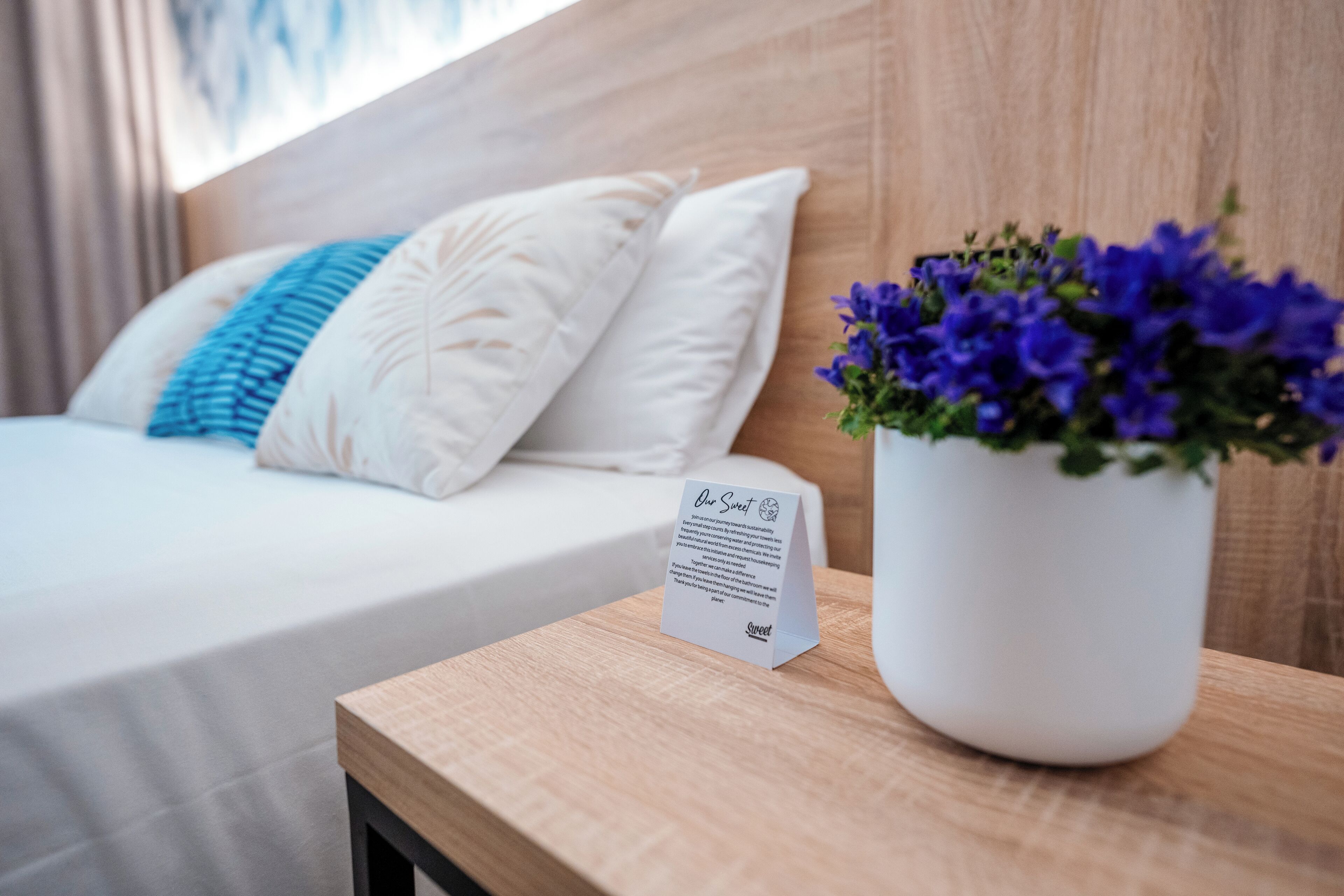 A neatly made bed with white linens and a blue pillow, a small white sign with text, and a white vase with purple flowers on a wooden bedside table.