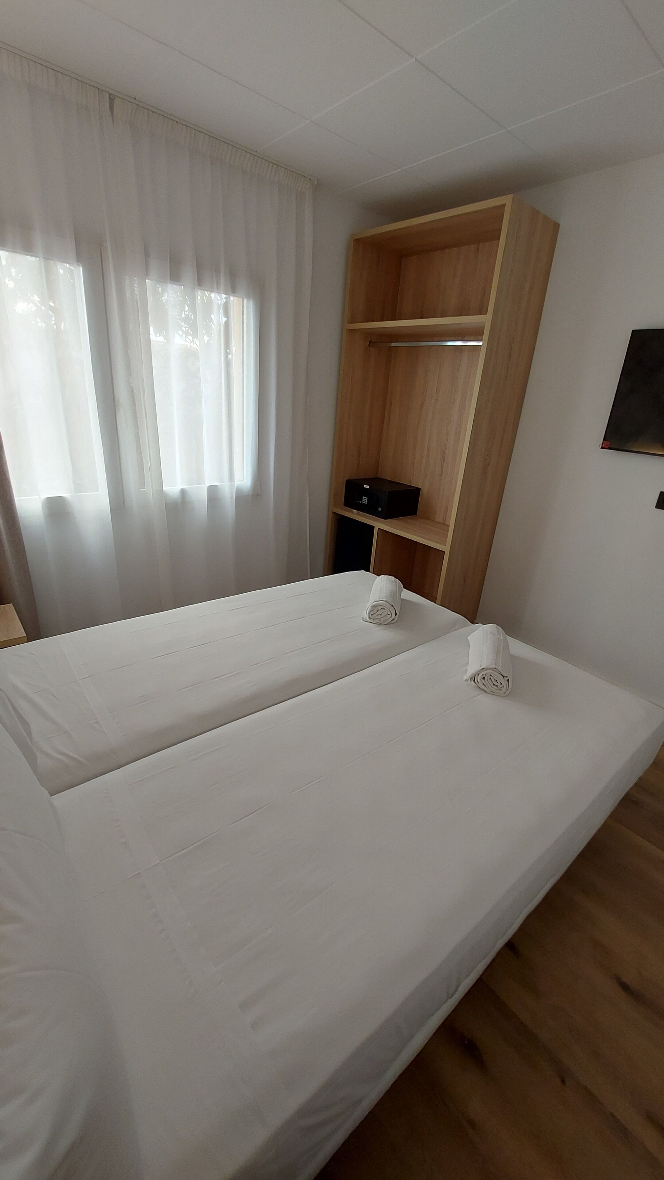A room with a single bed, a wooden wardrobe, a small shelf with a black object, and a window with sheer curtains.