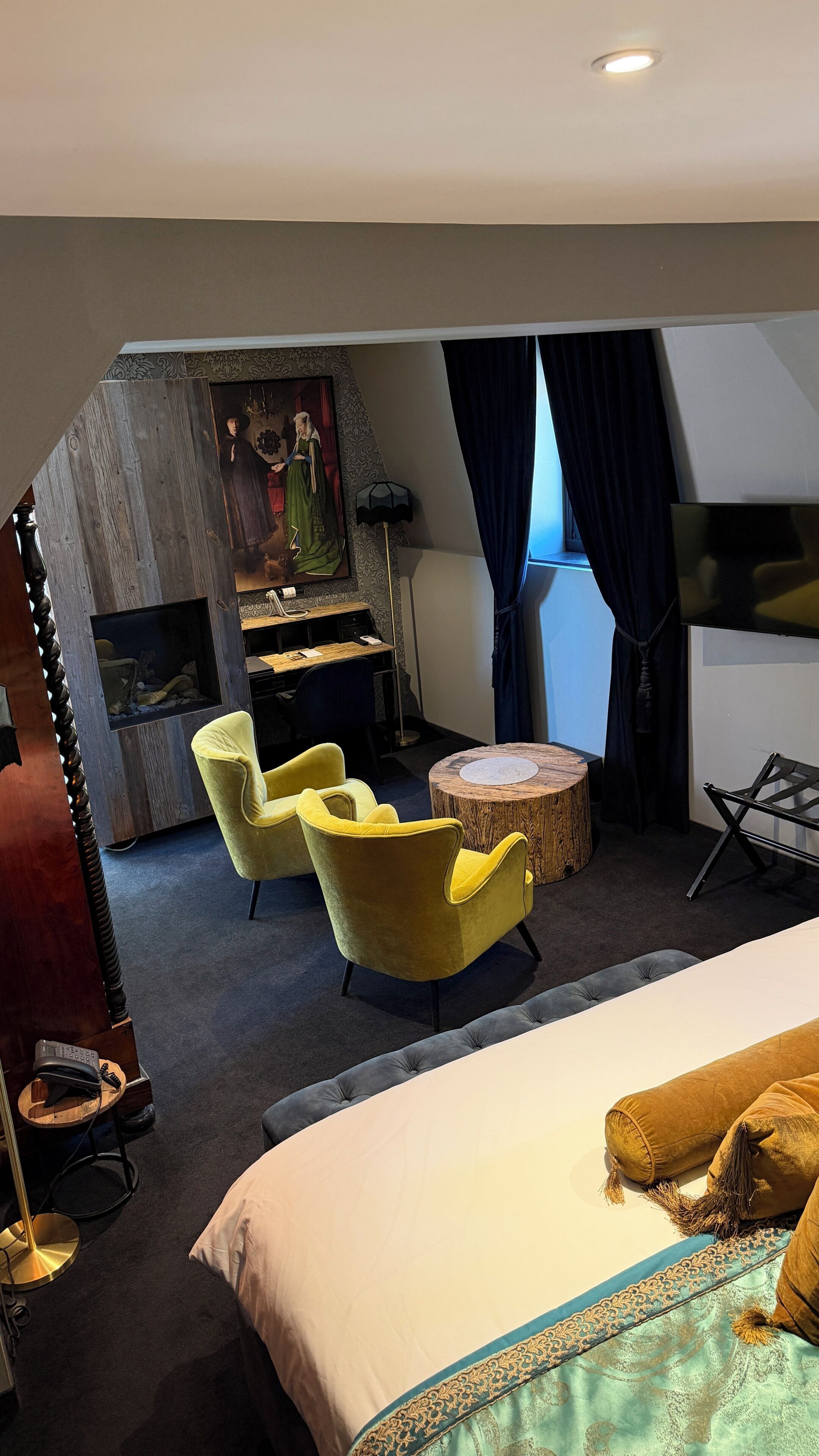 A hotel room with a bed, two yellow armchairs, a round wooden table, a desk with a computer, and a painting on the wall.