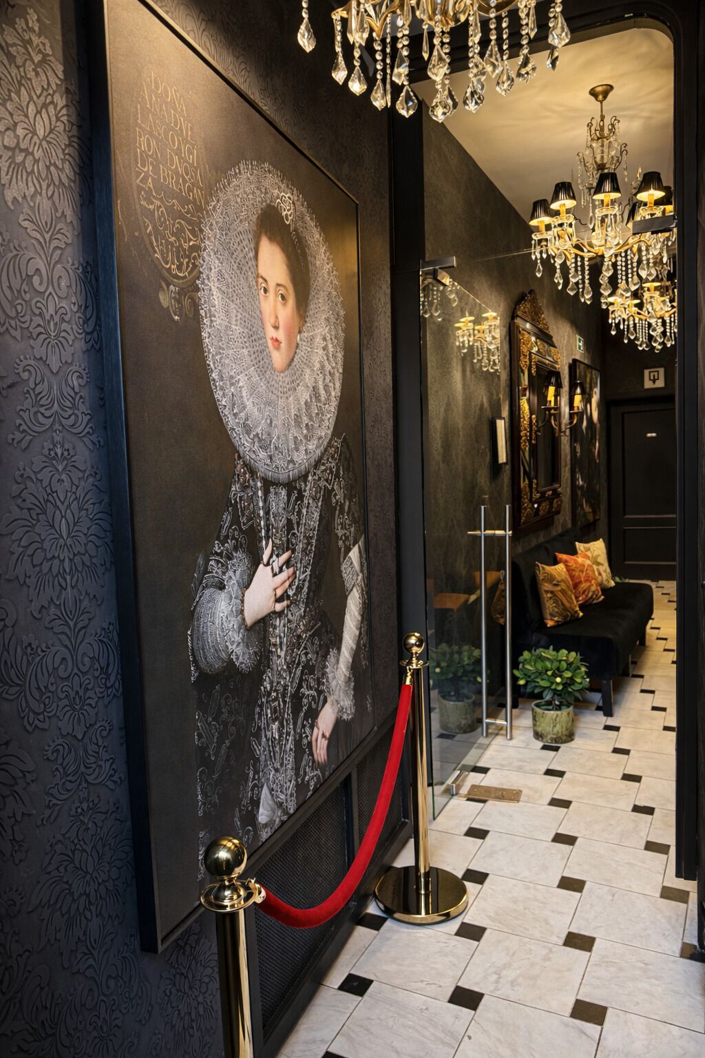 A hallway with a large portrait, a red velvet rope barrier, a chandelier, and a black sofa with cushions.
