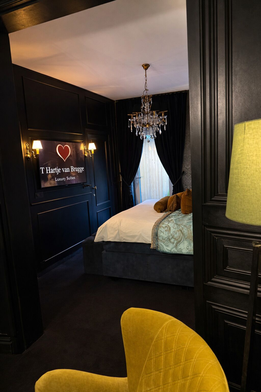 A hotel room with a bed, a chandelier, a sign reading 'T Hartje van Brugge Luxury Suites,' and a yellow chair.