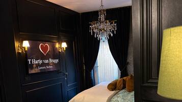 A hotel room with a bed, a chandelier, a sign reading 'T Hartje van Brugge Luxury Suites,' and a yellow chair.