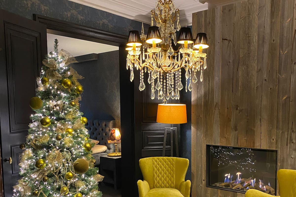 A living room with a Christmas tree, a chandelier, yellow armchairs, a fireplace, and a round wooden coffee table.