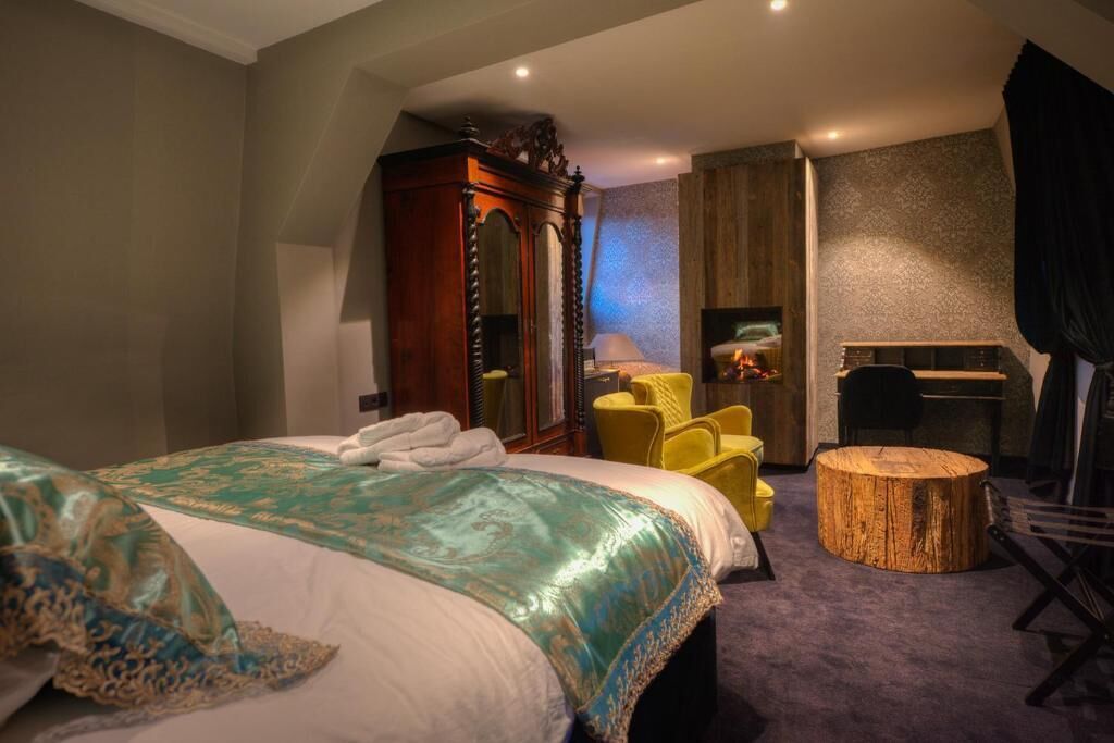A bedroom with a large bed, a wooden wardrobe, a yellow armchair, a round wooden stool, and a desk with a chair.