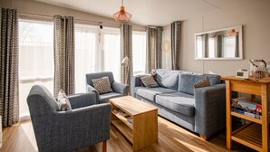 A living room with a sofa, armchair, coffee table, and a wooden side table with a clock and books. - Gemütliches Chalet im Ferienpark de Oase Renesse (Renesse)