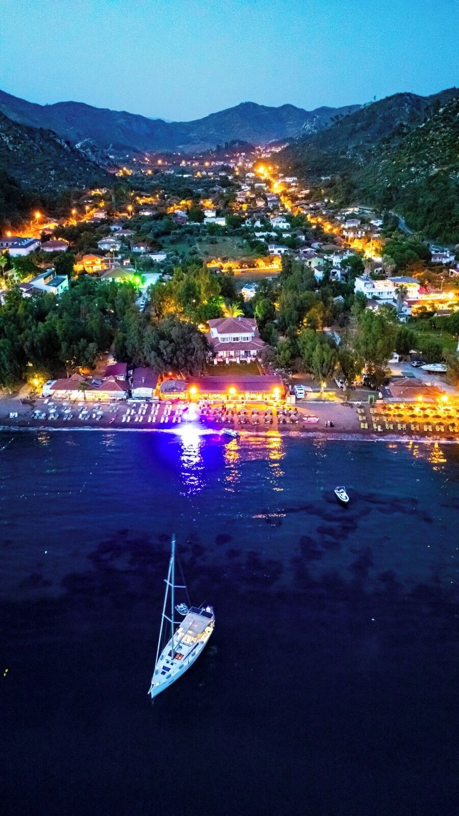 Vista aérea de un pueblo costero al anochecer, con un puerto deportivo, barcos e edificios iluminados.