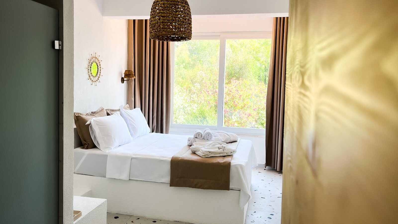 Un dormitorio con cama, lámpara de noche, ventana con cortinas y vistas al exterior.