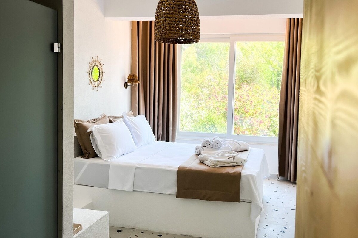 A bedroom with a bed, a bedside lamp, a window with curtains, and a view of greenery outside.