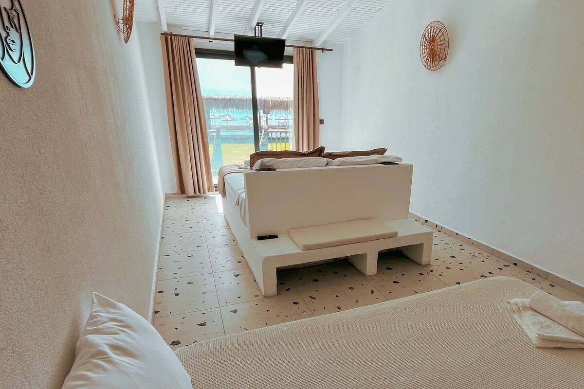 A room with a bed, a TV, a window with curtains, and a view of the beach.