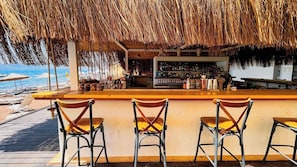 Un bar situé en bord de mer, doté d’un toit de chaume, d’un comptoir en bois et de chaises en métal.