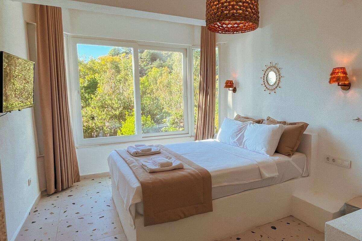 A bedroom with a bed, a window, a bedside table, a wall-mounted light, and a bathroom with a sink.