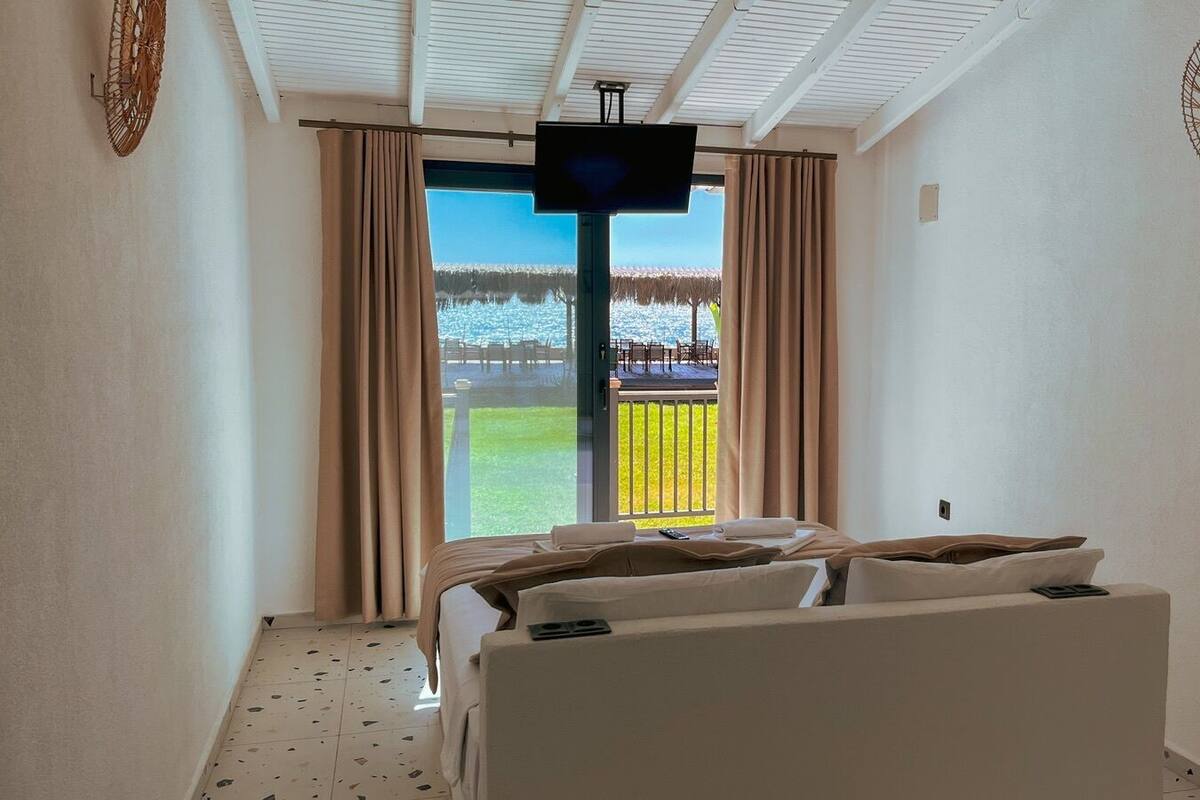 A hotel room with a bed, a TV, a balcony with a view of a beach and palm trees, and a ceiling fan.
