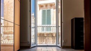 A room with a sliding glass door leading to a balcony with a metal railing and a view of a building with green shutters. - Casa Mila (Ostuni)