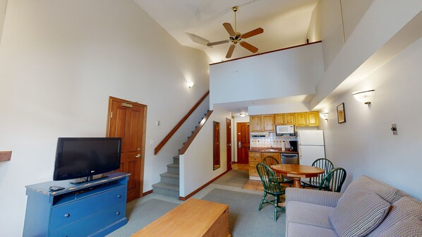 A living room with a TV, blue cabinet, wooden coffee table, green chairs, and a sofa. A staircase leads to a kitchen with wooden cabinets and a microwave. - Hearthstone Lodge #336 | 3 Peaks Rentals (Sun Peaks)