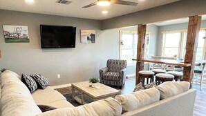 Un saló modern amb un sofà gran, una taula de centre i un butàc armchair amb dibuixos. Hi ha un televisor de pantalla plana muntat a la paret i una zona de menjador amb una taula de fusta i caires.