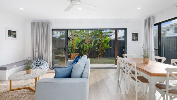 A modern living room with a sofa, a dining table with chairs, a small round table with a plant, and a sliding glass door leading to a garden. - Beach Pad on Nautilus (Kingscliff)