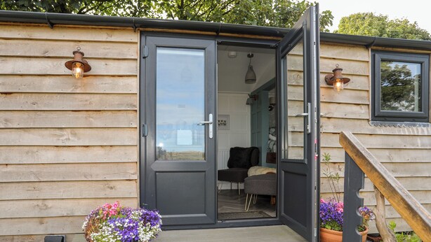 A wooden cabin with a dark grey door, a small porch with a bench, and two potted plants with purple flowers. - Shepherds Stone (Holyhead)