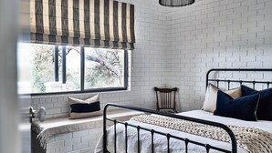Un dormitorio con pared de ladrillo blanco, una ventana con cortinas a rayas, una cama con estructura metálica y una silla de madera.