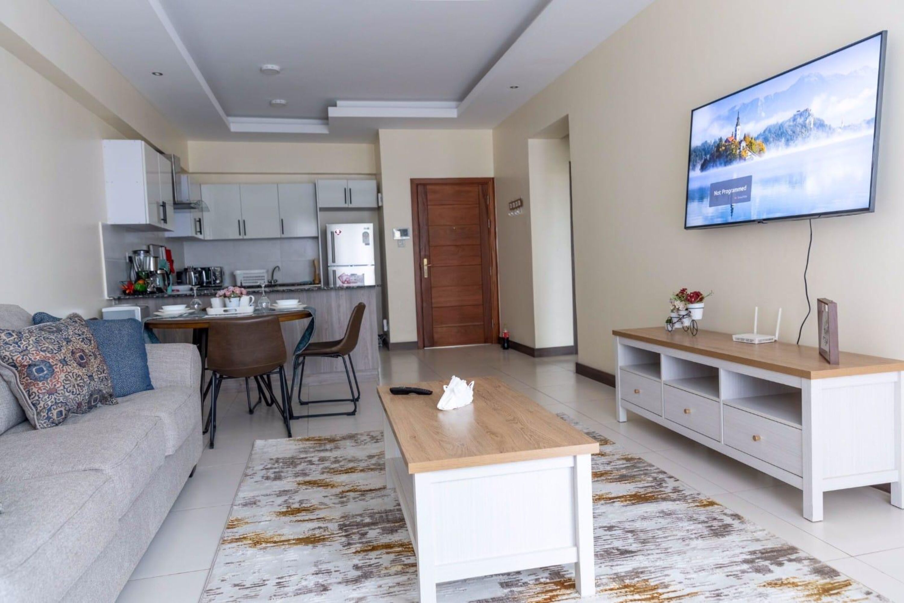 Una sala de estar moderna con un sofá, una mesa de centro y un televisor. Se ve una cocina al fondo con armarios y electrodomésticos.
