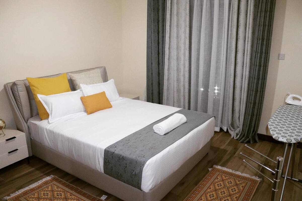 A neatly made bed with a grey bedspread, white and yellow pillows, and a rolled white towel. There is a small table with a patterned tablecloth and a chair with a metal frame. The room has wooden flooring and a patterned rug.