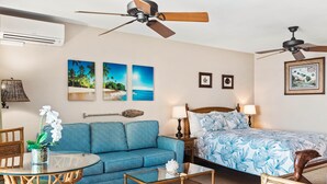 A room with a bed, a blue sofa, a glass coffee table, wicker chairs, and a ceiling fan.