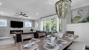 A modern dining area with a long table set for six, a fireplace, a flat-screen TV, and a large window with a view of greenery. - 13SC Beach, Private Pool, 10 foot movie theater screen (Hilton Head Island)