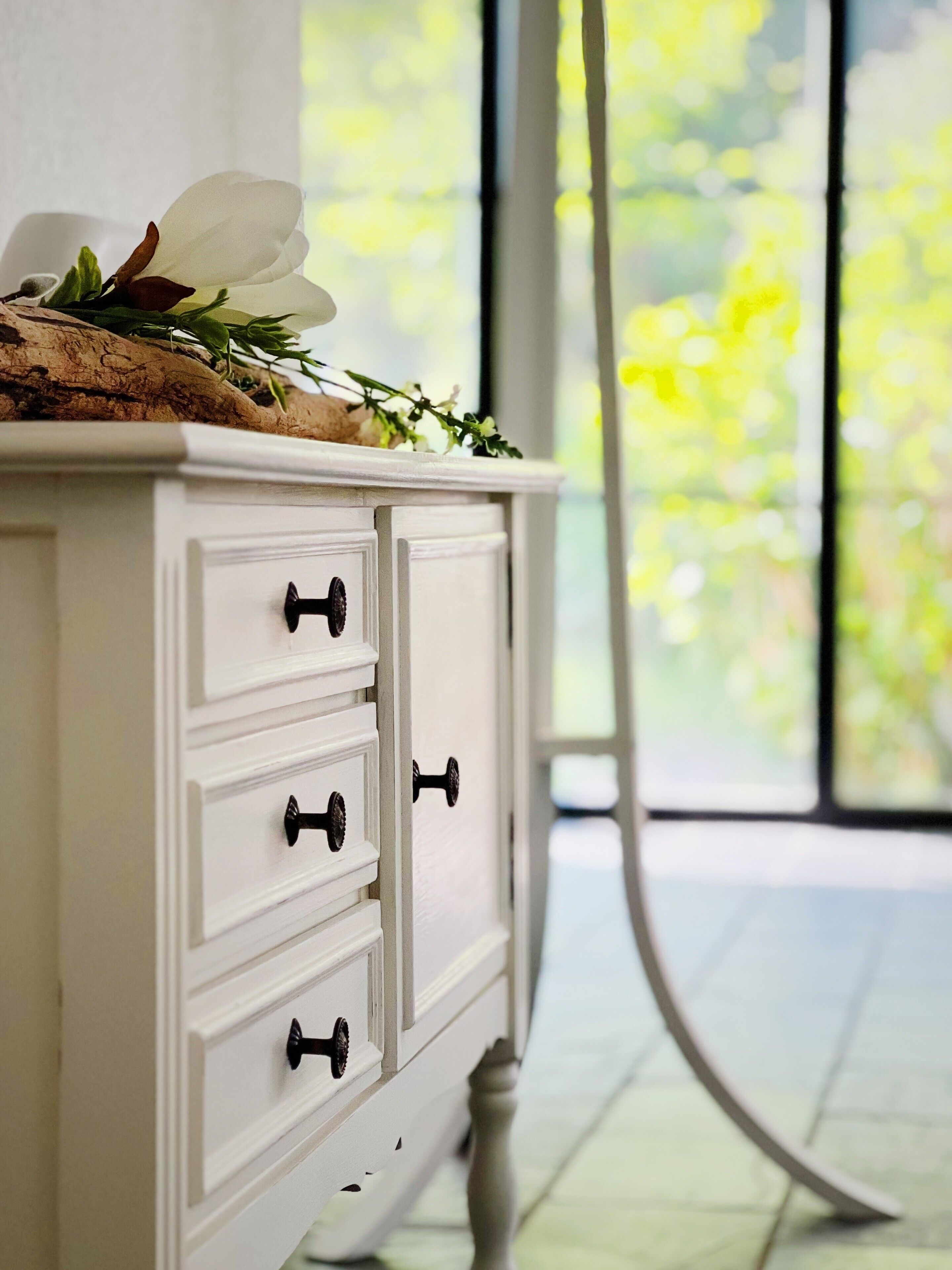 Een witte dressoir met zwarte knoppen, een bloemstuk erboven en uitzicht op groen door een raam.