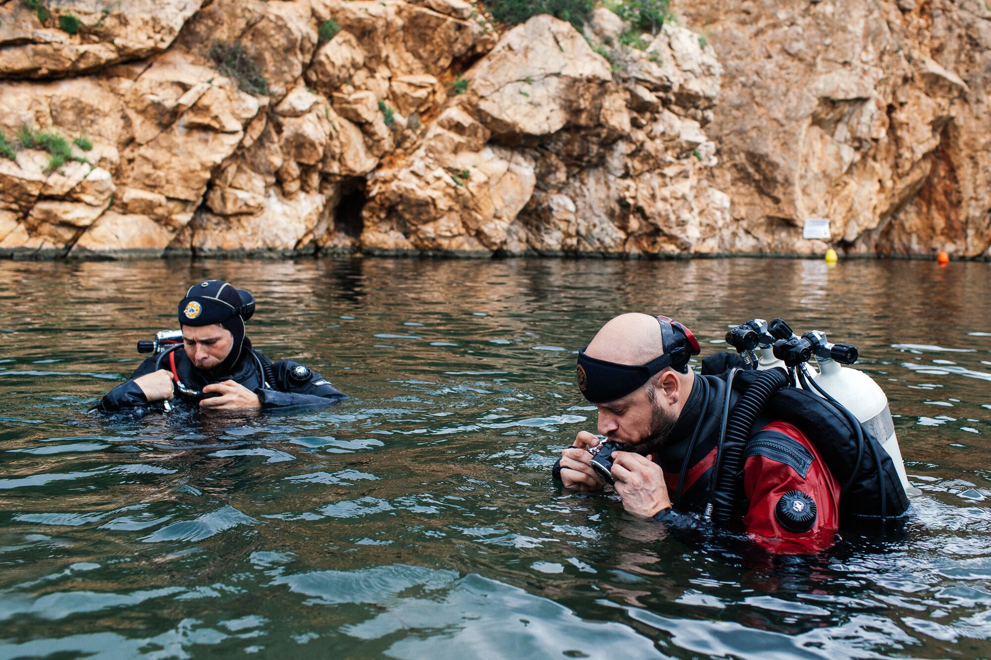 Two individuals in water with scuba gear, a rocky background, and a buoy.