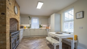 Una cocina con horno de ladrillo, armarios blancos, suelo con diseño espiga y una zona de comedor con banco y mesa.