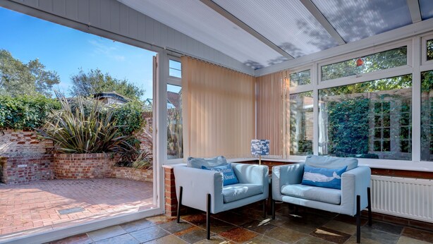 Un patio iluminado por el sol con dos sofás azul claro y una pequeña mesa, rodeado de amplias ventanas que dan a un jardín y edificios cercanos.