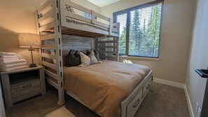 Une chambre avec un lit superposé, une table de chevet avec une lampe, une fenêtre avec des stores et un sol recouvert d’une moquette.
