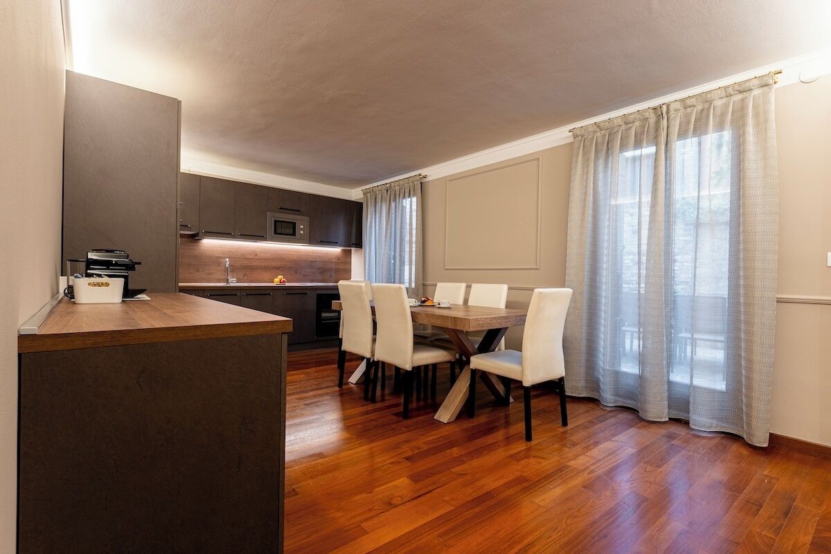 A modern kitchen with wooden flooring, a dining table with chairs, and a large window with curtains.