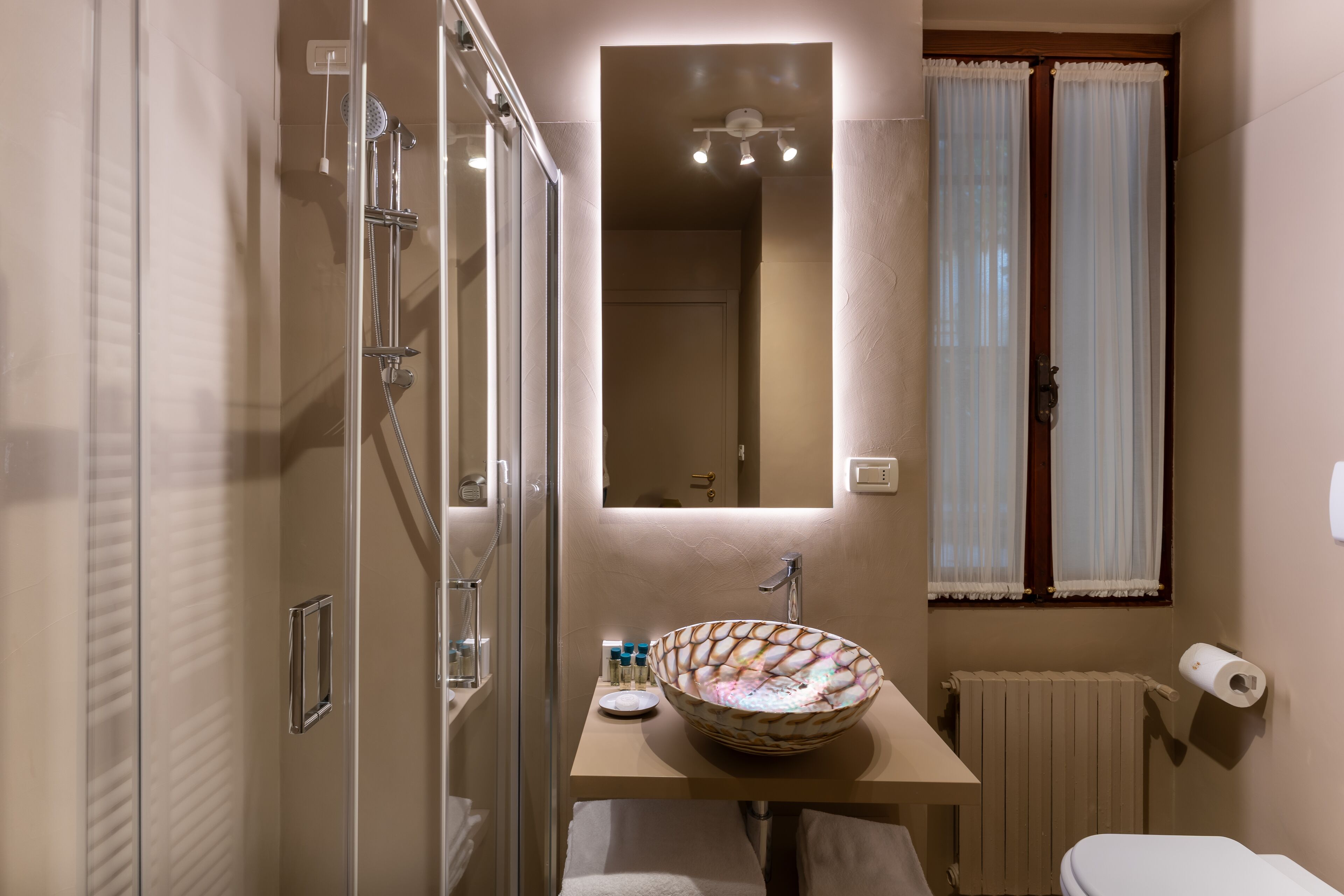 A bathroom with a glass shower enclosure, a sink with a decorative bowl, a mirror above the sink, a toilet, and a window with curtains.