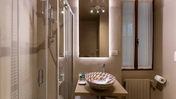A bathroom with a glass shower enclosure, a sink with a decorative bowl, a mirror above the sink, a toilet, and a window with curtains.