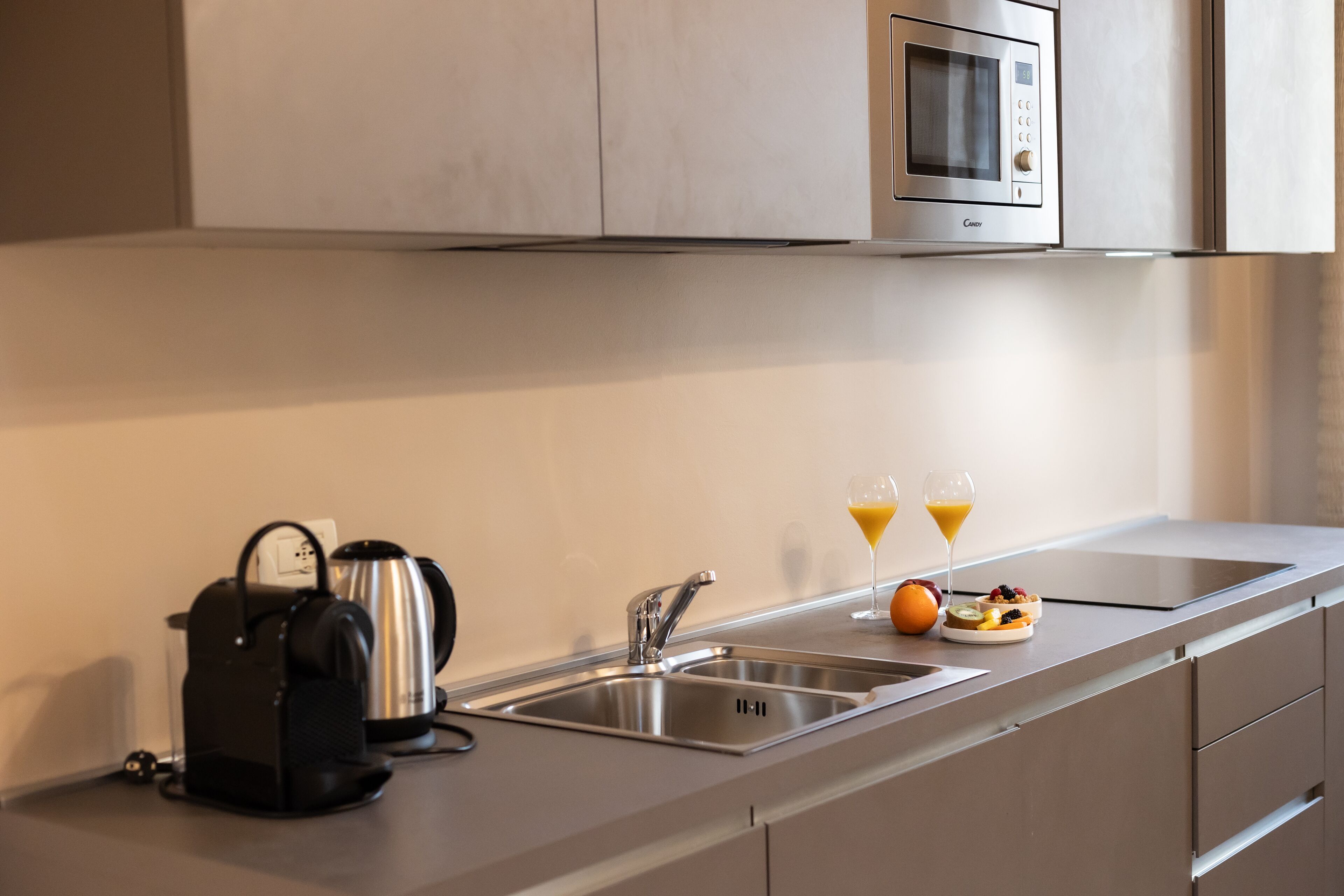 Una cocina moderna con microondas integrado, tetera, lavabo y frutero con vasos de jugo de naranja.