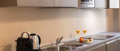 Una cocina moderna con microondas integrado, tetera, lavabo y frutero con vasos de jugo de naranja.