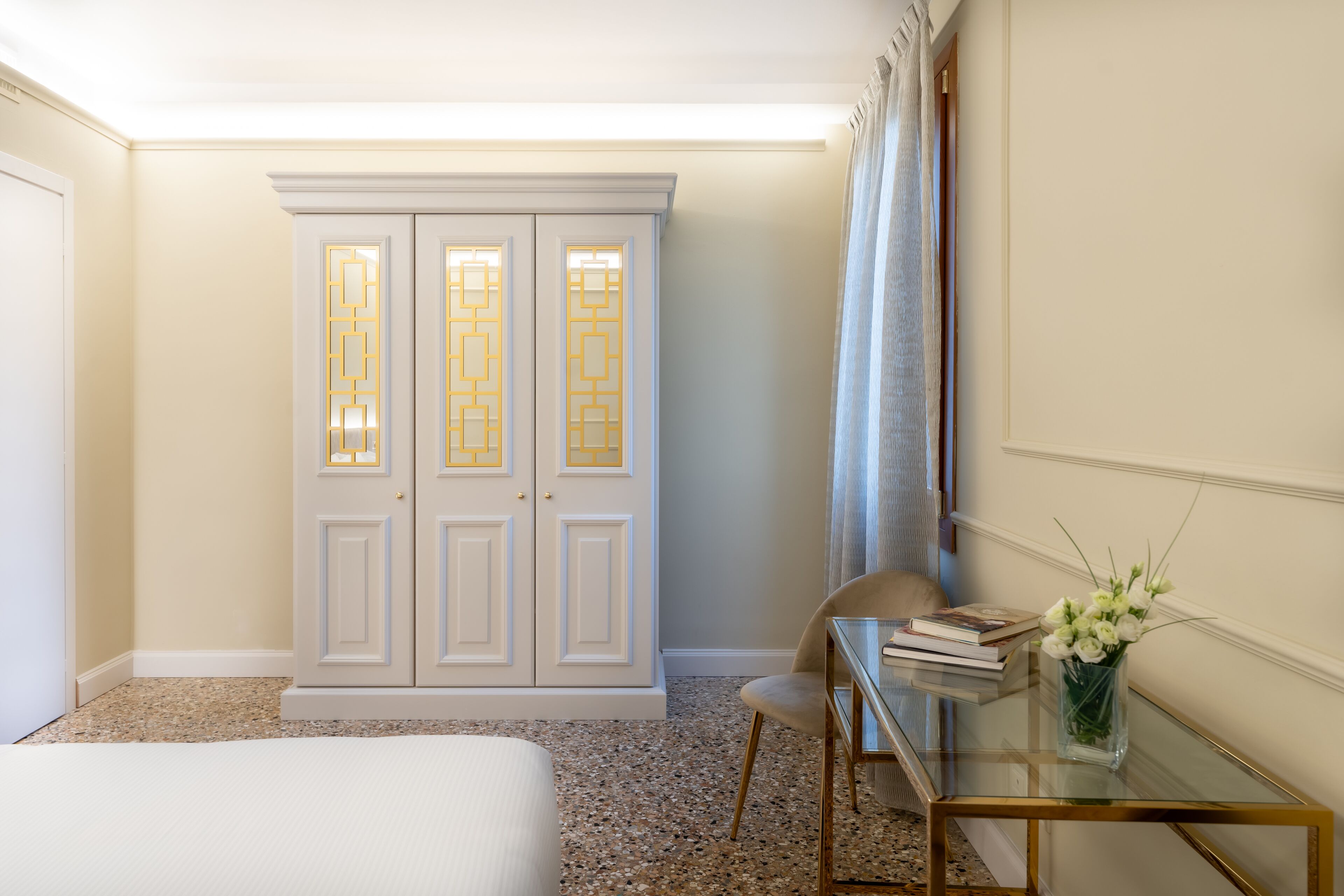 A hotel room with a bed, a glass table with a vase of flowers, a chair, and a wardrobe with decorative panels.